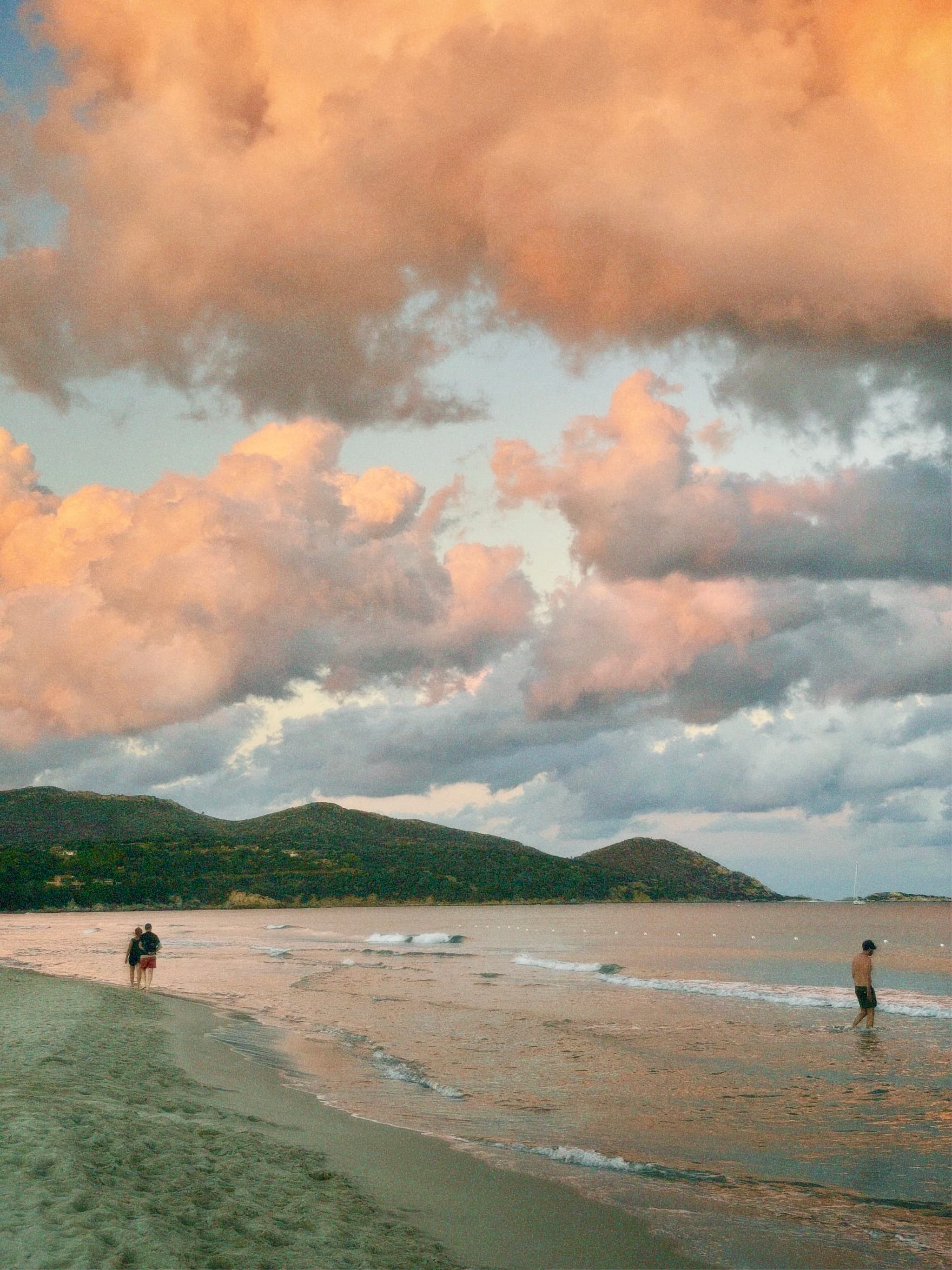Two beachgoers walk along a serene shoreline under a vast, colorful sky with dramatic clouds at sunset