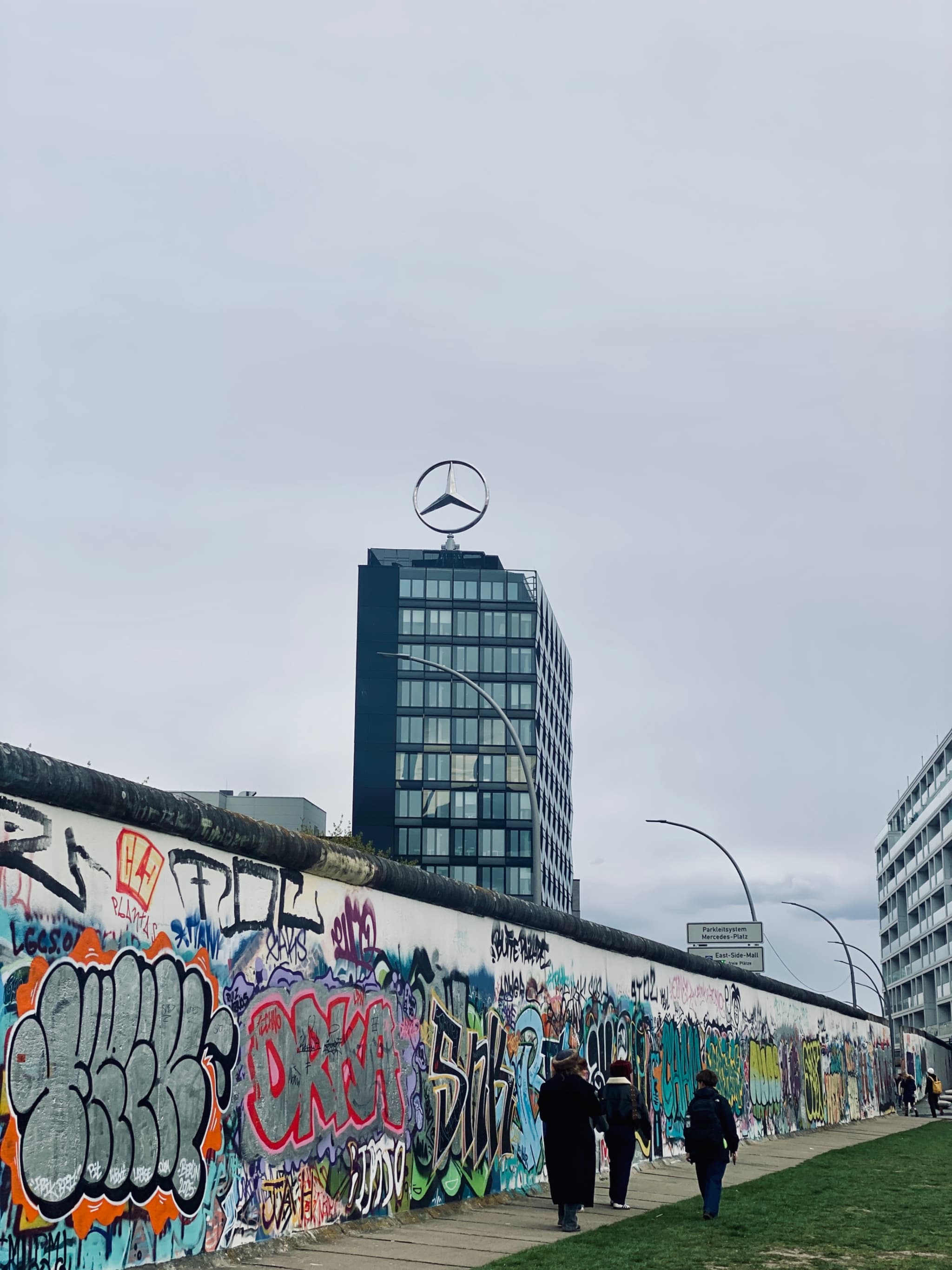 Colorful graffiti adorns the Berlin Wall, with the Mercedes-Benz Tower looming in the background under a gray, overcast sky, evoking a gritty yet vibrant urban aesthetic
