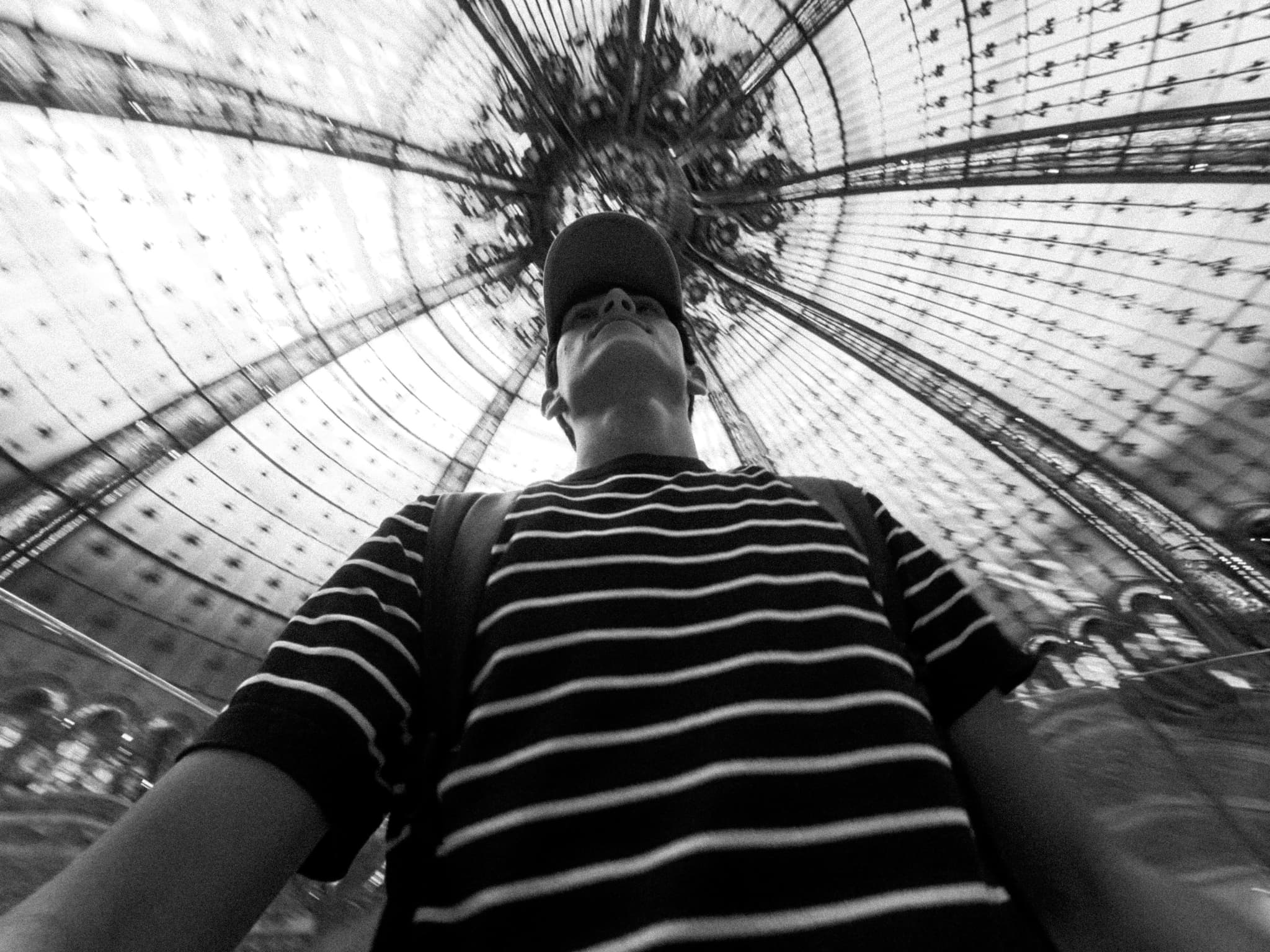 A person in a striped shirt is captured from below, looking up through a glass ceiling with intricate metalwork, under moody natural light