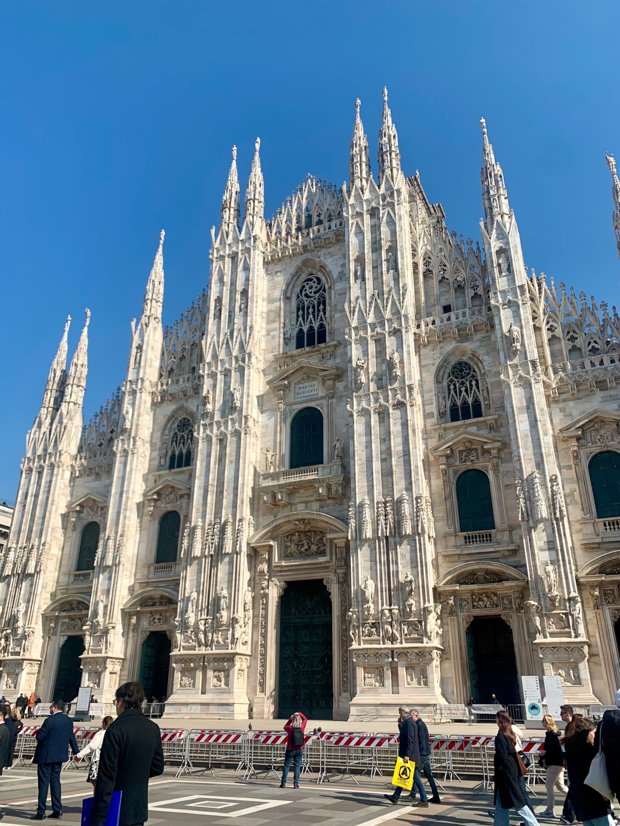 Sunlit view of the majestic Milan Cathedral, bustling with people, set against a clear blue sky, capturing its grand Gothic architecture