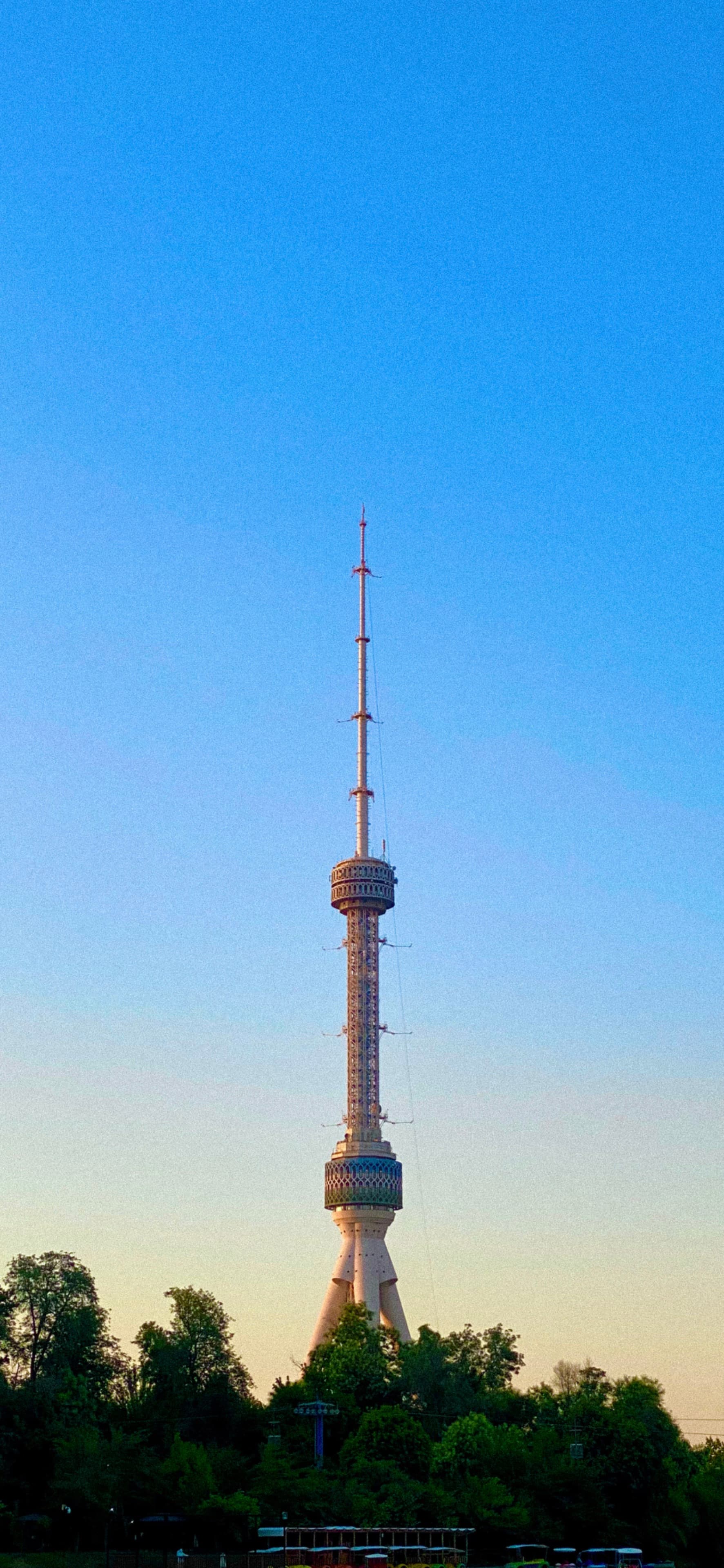 A modern, tall TV tower stands against a clear blue sky, framed by lush green trees, suggesting a serene, urban setting
