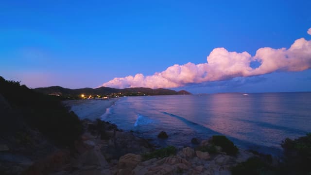 A tranquil coastal sunset scene with soft clouds, calm waters, and gentle shoreline under a deep blue sky