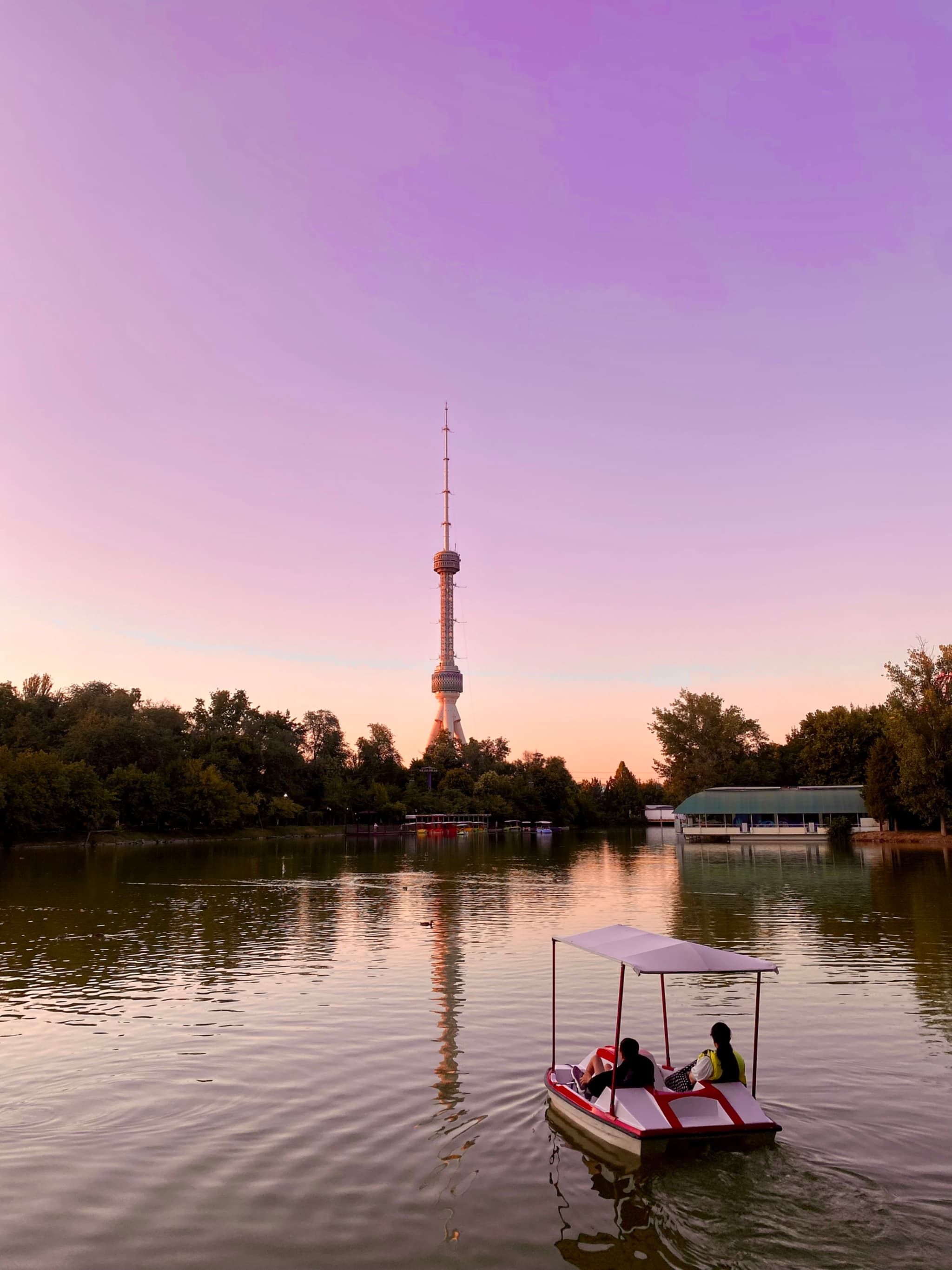 A serene sunset scene by a lake, with a modern tower reflecting in the water and a small boat with people enjoying the tranquil view