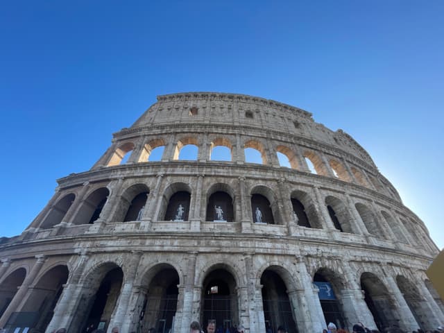 Amidst a clear blue sky, the majestic Roman Colosseum stands tall against the backdrop, bathed in warm sunlight, highlighting its intricate arches and historical grandeur