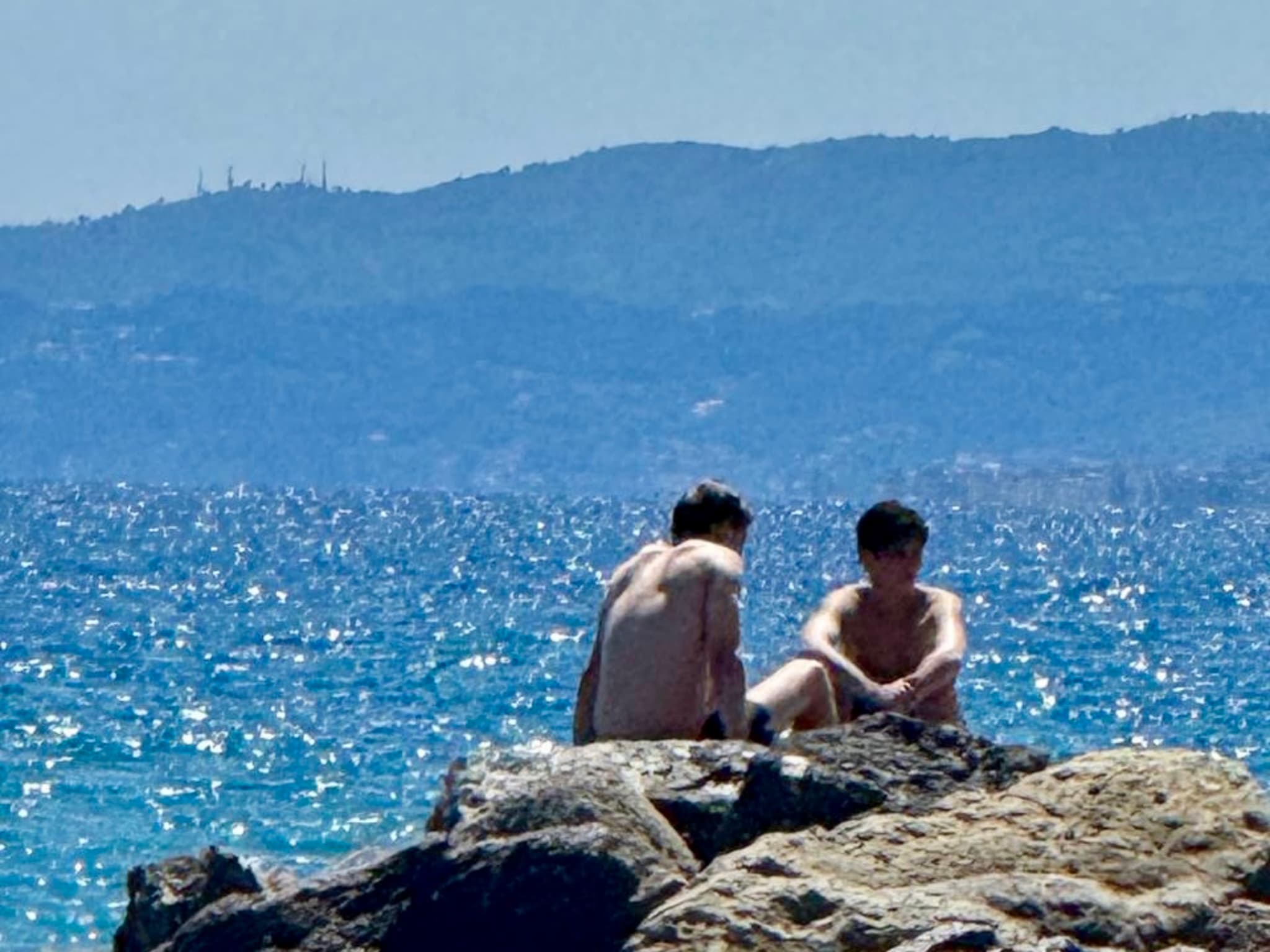 Two boys relax on a rocky beach, gazing out at sparkling blue waters under a bright, sunny sky