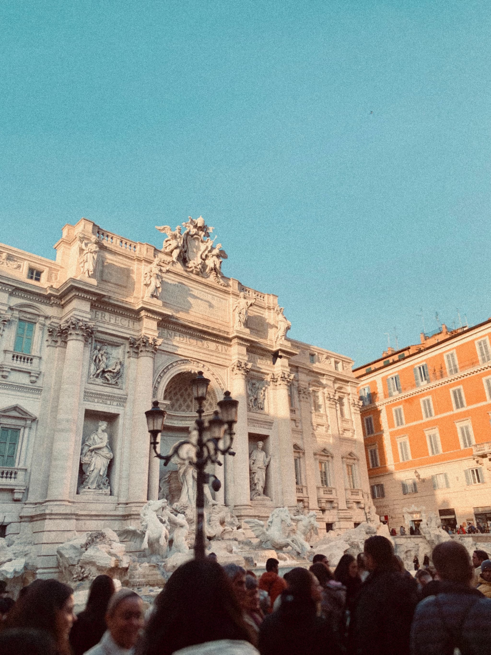 Sunlit Trevi Fountain in Rome, Italy, with crowds in the foreground, clear blue sky, and warm tones
