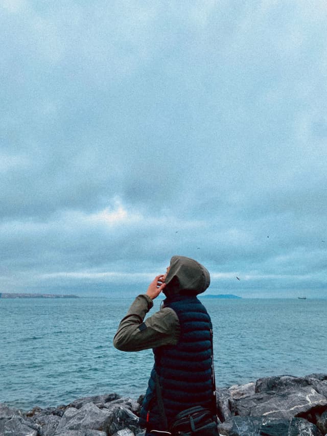 A man gazes at a vast, overcast sky above a serene coastal scene, with muted colors and a somber, contemplative mood