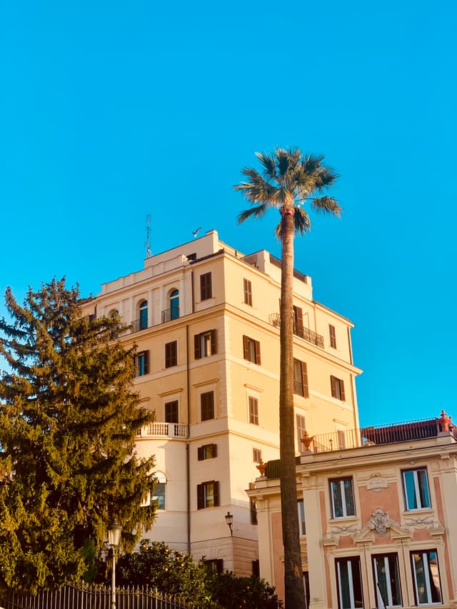 A classic Italian building stands under a bright blue sky, with a palm tree in the foreground, evoking a warm, sunny atmosphere
